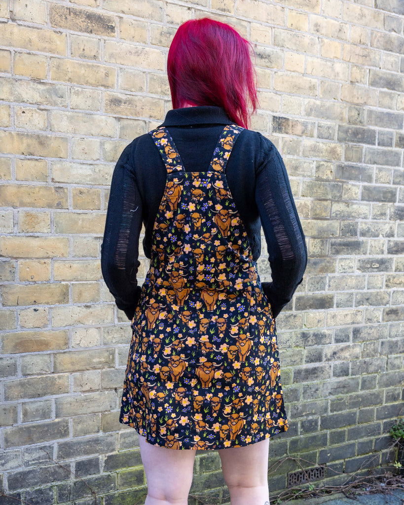 Florence, a femme model with red hair and tattoos, is stood outside in a mews in Hove wearing Highland Cows Stretch Twill Pinafore Dress with a long sleeved black top underneath and black boots. The black pinafore dress has an all over print of Highland cows amongst purple flowers and grass designed by Jen James. Florence is stood with her back to the camera and hands by her sides.