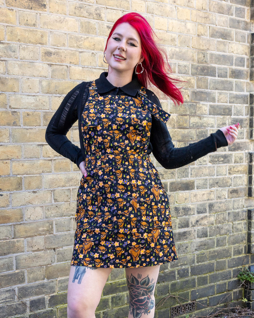 Florence, a femme model with red hair and tattoos, is stood outside in a mews in Hove wearing Highland Cows Stretch Twill Pinafore Dress with a long sleeved black top underneath and black boots. The black pinafore dress has an all over print of Highland cows amongst purple flowers and grass designed by Jen James. Florence is facing the camera smiling with one hand on her hip and other swishing her hair.