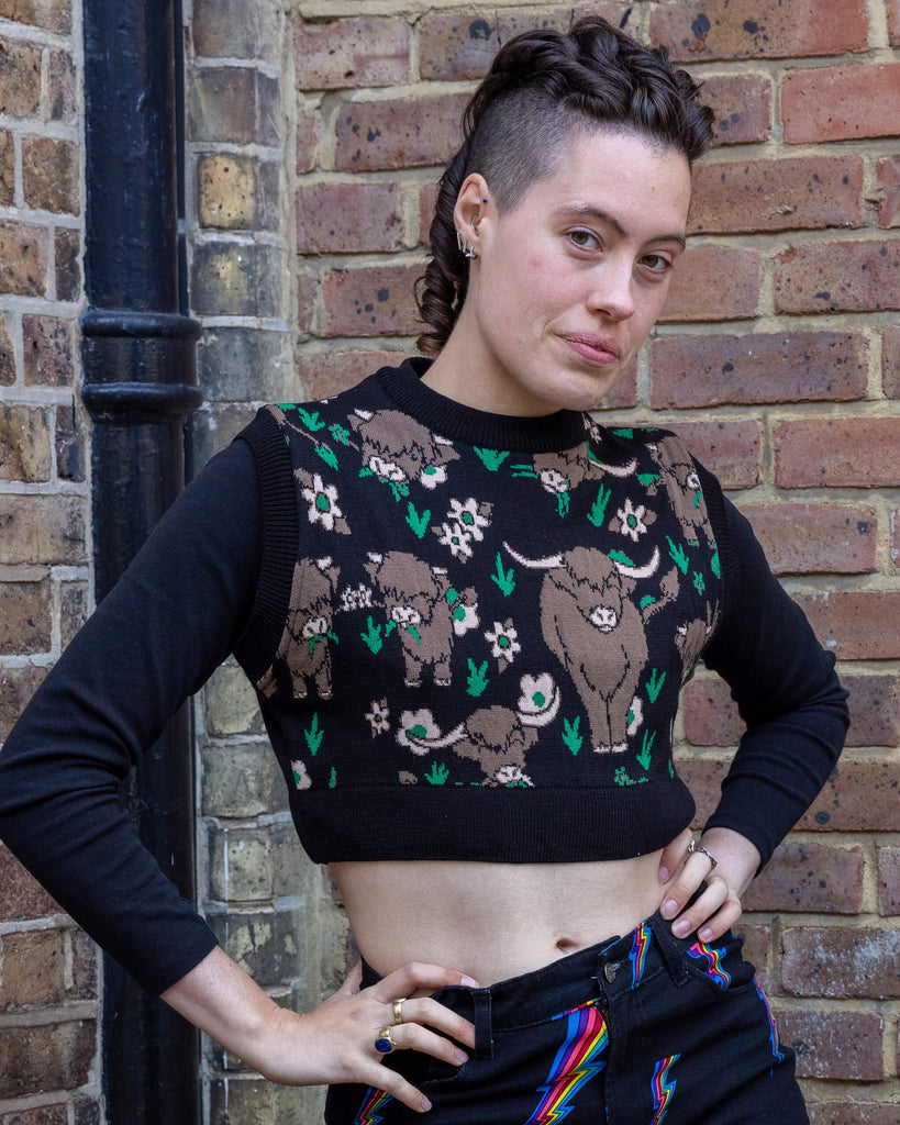 Close-up of a person modelling a black cropped knitted tank top with a highland cows print. The design features brown cows and floral details in green and pink. Paired with black jeans featuring rainbow lightning bolts. Run & Fly creates playful, ethically made clothing that promotes slow fashion.