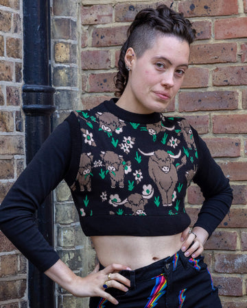 Close-up of a person modelling a black cropped knitted tank top with a highland cows print. The design features brown cows and floral details in green and pink. Paired with black jeans featuring rainbow lightning bolts. Run & Fly creates playful, ethically made clothing that promotes slow fashion.
