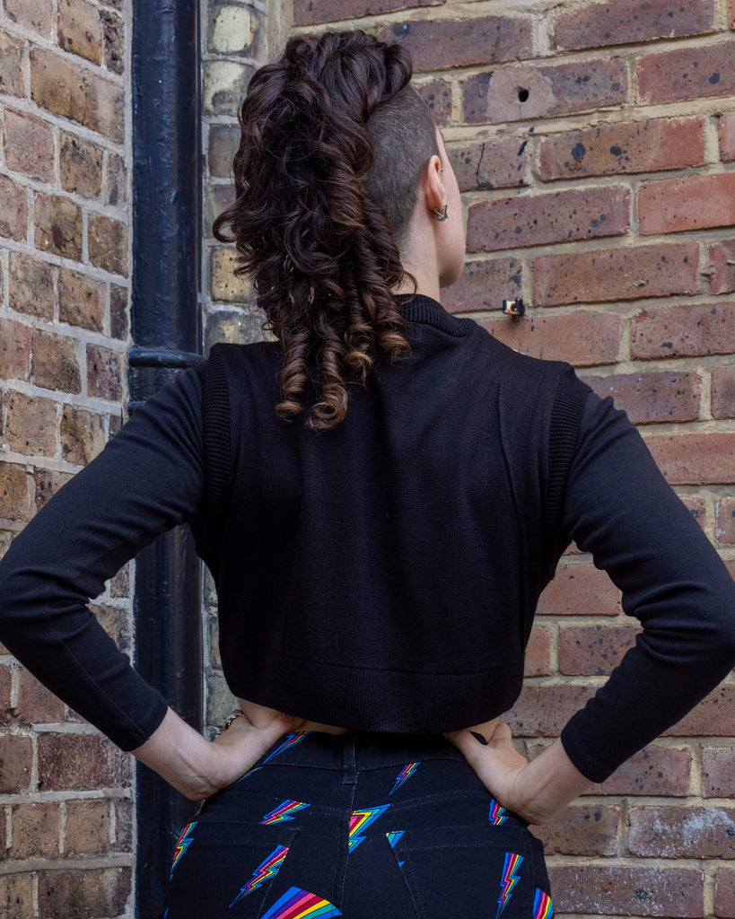 Back view of a person wearing a black cropped knitted tank top from Run & Fly, paired with black jeans. The top is plain at the back with long sleeves underneath, showing off its cropped design. Run & Fly is a small ethical fashion brand offering unique and sustainable clothing choices.