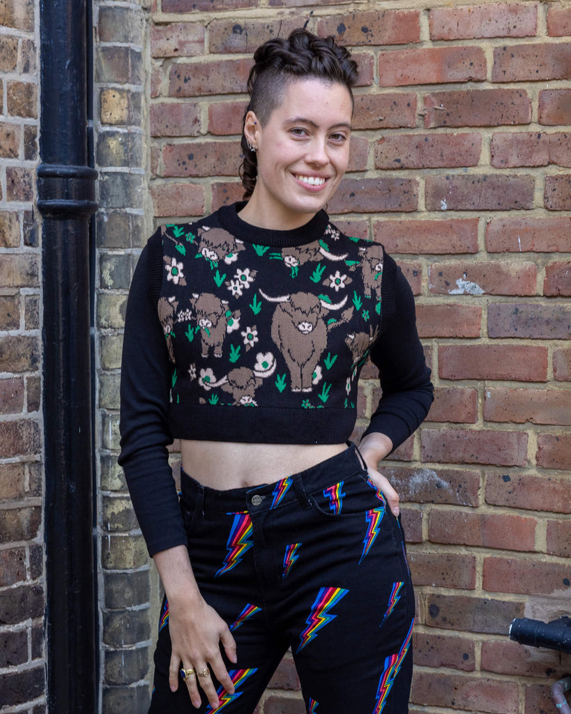 A person wearing a black cropped knitted tank top featuring a cute highland cows print, paired with black jeans adorned with rainbow lightning bolts. The top showcases brown highland cows surrounded by green grass and pink flowers. Run & Fly is a small ethical slow fashion brand known for quirky and sustainable knitwear designs.