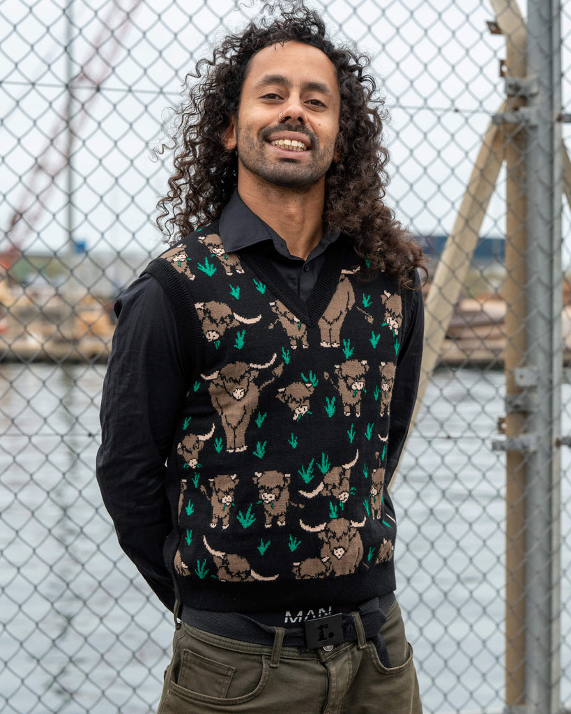 Model Rich wears the Run & Fly Highland Cow Tank Vest made in England, featuring a brown highland cow pattern by Jen James Design on a black knit. Styled over a black shirt with khaki trousers for a unisex look.