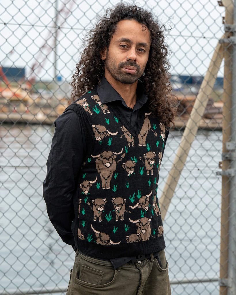 Model Rich poses wearing the Run & Fly Highland Cow Tank Vest, a unisex black knit made in England with an illustrated highland cow pattern by Jen James Design.
