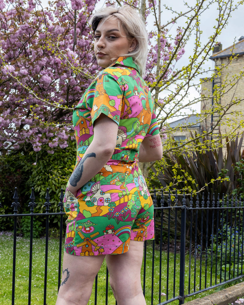 The Run & Fly X The Mushroom Babes Hills Stretch Twill Playsuit worn by a femme tattooed model with split blonde and black hair. She is stood outside on grass underneath a blossom tree, she is facing the side and looking over her shoulder at the camera with her hands in her pockets. . The stretch twill playsuit print features green, pink and orange hills with trees, mushroom cottages, ponds, leading pathways and picnics.