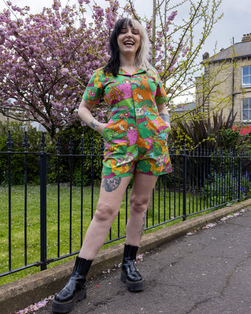 The Run & Fly X The Mushroom Babes Hills Stretch Twill Playsuit worn by a femme tattooed model with split blonde and black hair with black boots. She is stood outside on grass underneath a blossom tree, she is facing forward and smiling with her hands in the playsuit pockets. The stretch twill playsuit print features green, pink and orange hills with trees, mushroom cottages, ponds, leading pathways and picnics.