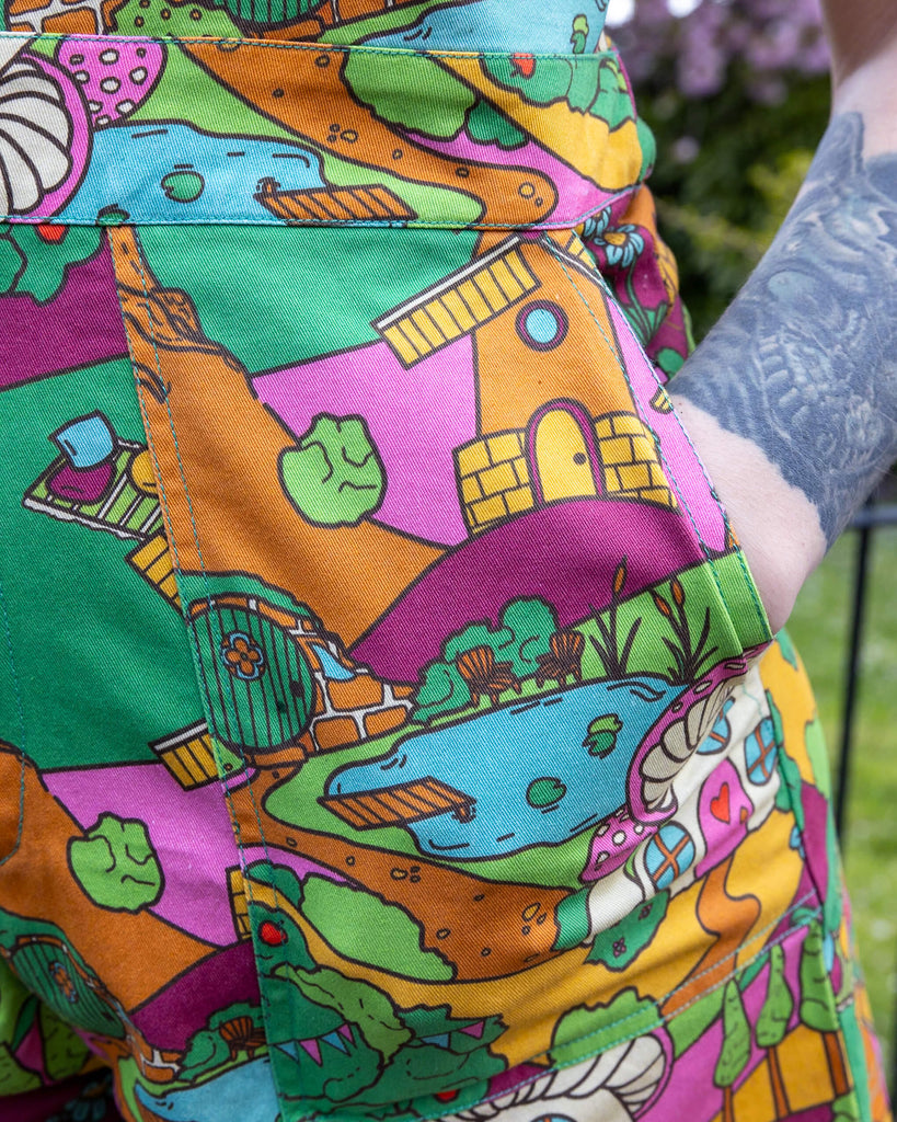 Close up of the Run & Fly X The Mushroom Babes Hills Stretch Twill Playsuit worn by a femme tattooed model. She is stood outside on grass underneath a blossom tree with the close up showing her hand in the side pocket and the zip up middle. The stretch twill playsuit print features green, pink and orange hills with trees, mushroom cottages, ponds, leading pathways and picnics.