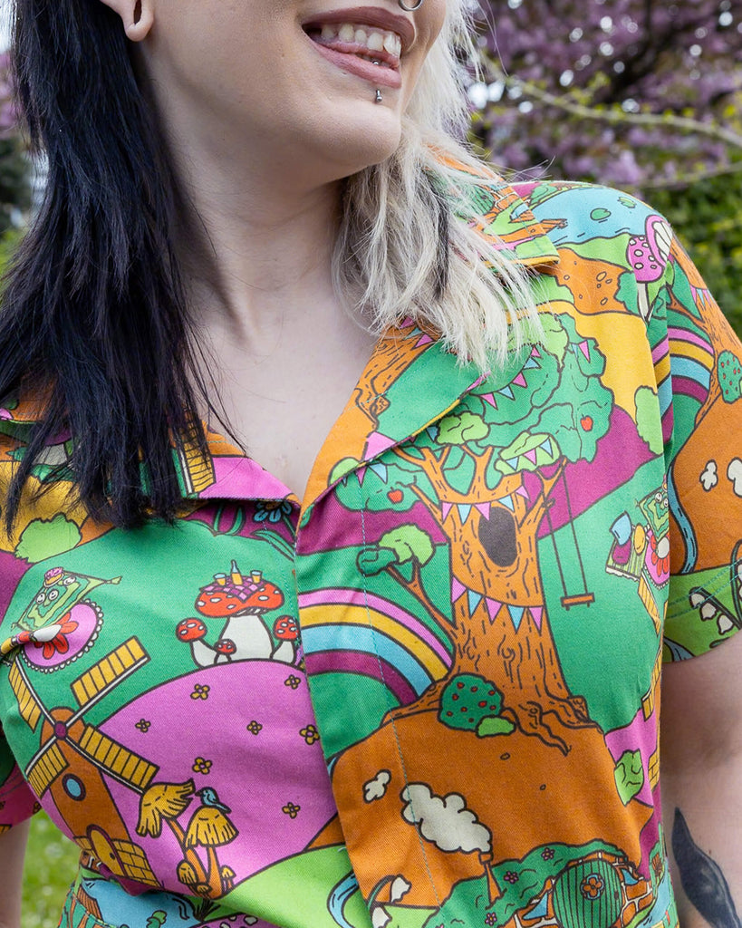 Close up of the Run & Fly X The Mushroom Babes Hills Stretch Twill Playsuit worn by a femme tattooed model with split blonde and black hair. She is smiling stood outside on grass underneath a blossom tree with the close up showing the collar top and zip detailing. The stretch twill playsuit print features green, pink and orange hills with trees, mushroom cottages, ponds, leading pathways and picnics.
