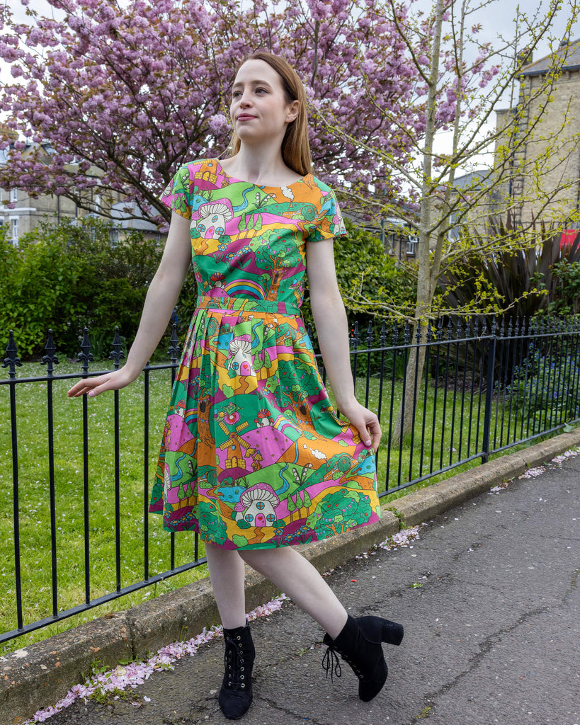 A woman in a colourful, mushroom-themed tea dress by Run & Fly poses outdoors, playfully lifting part of the skirt. The dress, cinched with a belt, features rainbows, hills, and fantasy creatures in vibrant hues of green, orange, and pink. She is standing in front of a blooming tree with soft pink flowers, her black ankle boots adding a chic touch to the look.