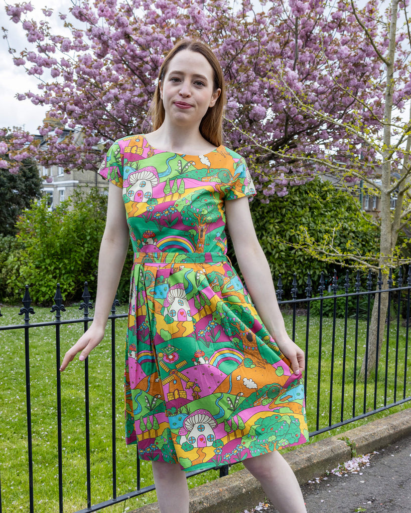 A smiling woman wearing a vibrant Run & Fly Mushroom Babes Hills belted tea dress stands confidently in a park. The colourful dress, adorned with rainbows, mushrooms, and hills, cinches at the waist with a belt, creating a flattering silhouette. The background showcases a lush green lawn and a blossoming pink tree, enhancing the whimsical theme of the outfit.