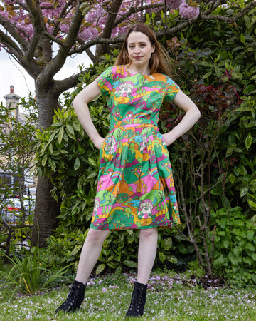 A woman strikes a confident pose outdoors in a whimsical Run & Fly Mushroom Babes Hills stretch belted tea dress. The knee-length dress showcases a colourful pattern of mushrooms, hills, and rainbows, in shades of green, orange, and pink. She stands on a grassy lawn surrounded by lush greenery and blossoming trees, exuding playful charm with her hands on her hips.