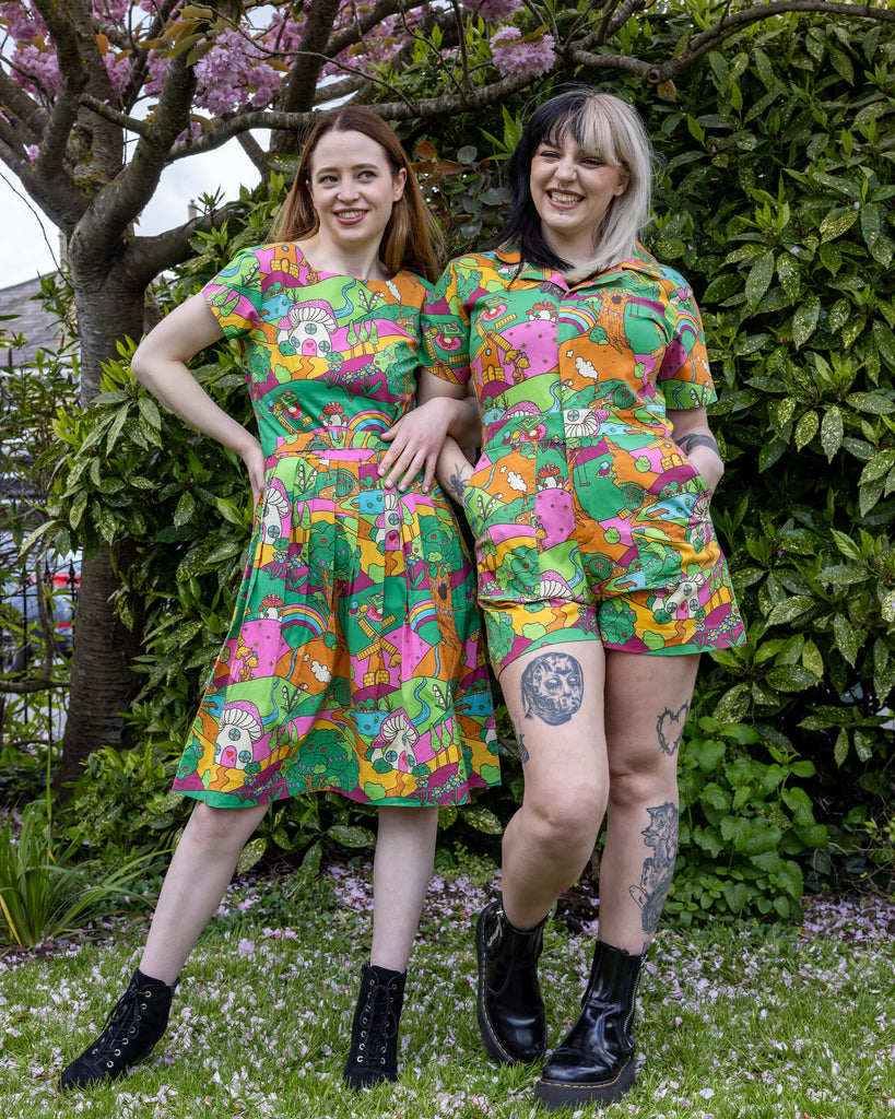 Two women pose side by side, each wearing the Mushroom Babes print by Run & Fly. One is in the belted tea dress and the other in a short-sleeved playsuit. The vibrant print features mushrooms, rainbows, and hills in bright, playful colours. They smile confidently, with a mix of black boots and tattoos adding an edgy touch to the whimsical outfits.