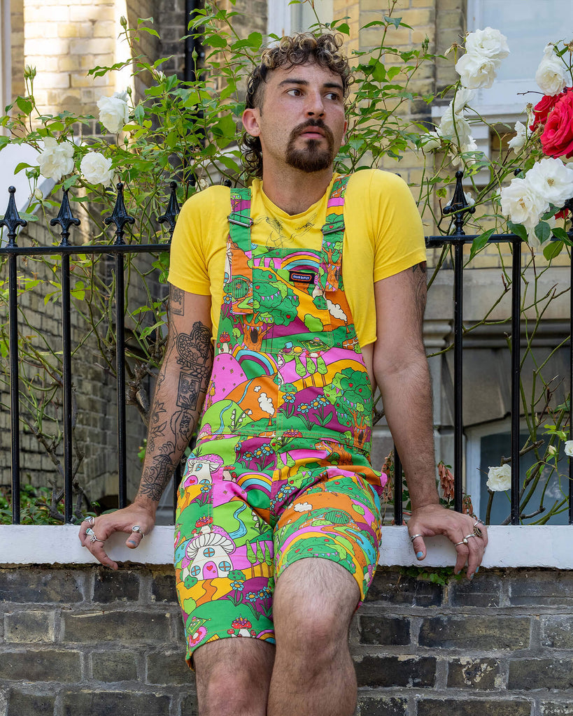 The Run & Fly x The Mushroom Babes Hills Stretch Twill Dungaree Shorts worn by a masc non binary person with a curly brown mullet haircut, a yellow crop top and white trainers. They are sat outside in front of a black railing with foliage and rose bushes behind. They are facing forward leaning back on the railing looking off to the right. The dungashorts are green, pink and orange hills with mushroom houses, flowers, rainbows and trees.