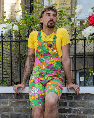 The Run & Fly x The Mushroom Babes Hills Stretch Twill Dungaree Shorts worn by a masc non binary person with a curly brown mullet haircut, a yellow crop top and white trainers. They are sat outside in front of a black railing with foliage and rose bushes behind. They are facing forward leaning back on the railing looking off to the right. The dungashorts are green, pink and orange hills with mushroom houses, flowers, rainbows and trees.