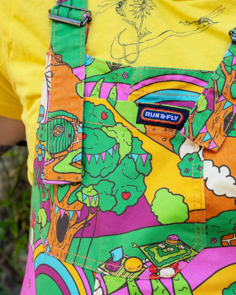Close up of the Run & Fly x The Mushroom Babes Hills Stretch Twill Dungaree Shorts worn by a masc non binary person with a yellow crop top. They are stood outside in front of foliage. The dungashorts are green, pink and orange hills with mushroom houses, flowers, rainbows and trees.