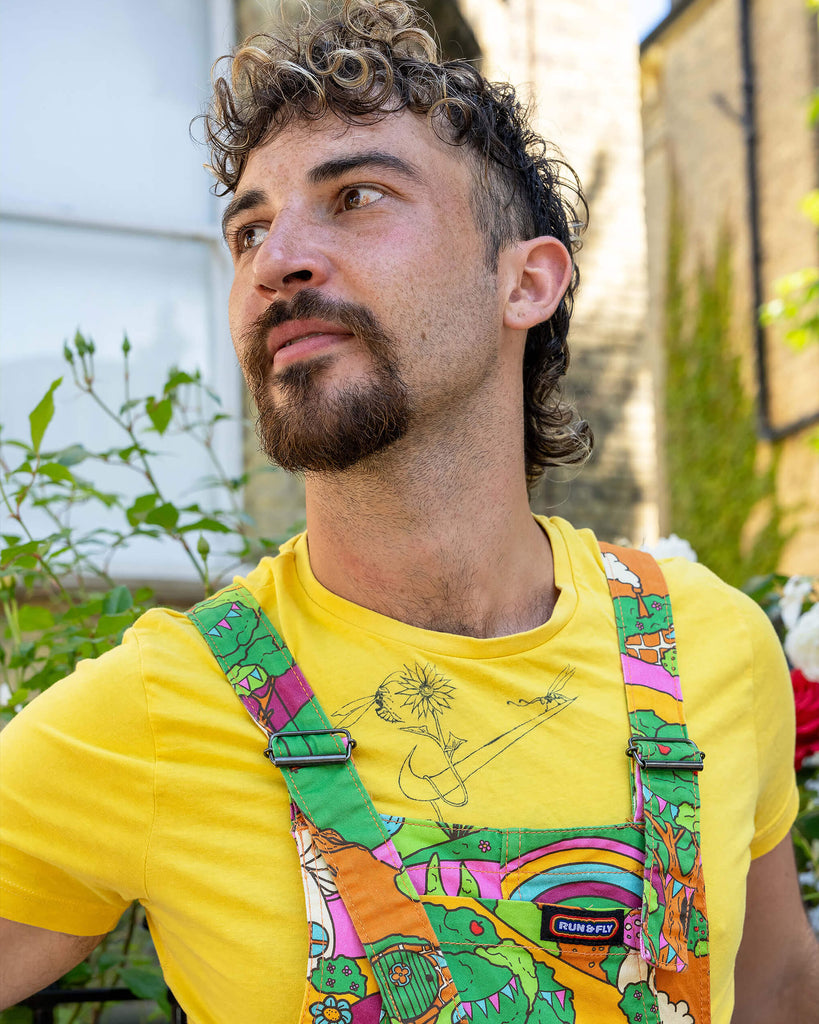 Close up of the Run & Fly x The Mushroom Babes Hills Stretch Twill Dungaree Shorts worn by a masc non binary person with a curly brown mullet haircut and a yellow crop top. They are stood outside in front of a black railing with foliage and rose bushes behind. They are facing forward smiling looking left. The dungashorts are green, pink and orange hills with mushroom houses, flowers, rainbows and trees.
