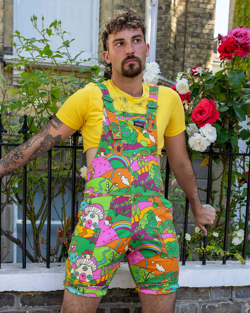 The Run & Fly x The Mushroom Babes Hills Stretch Twill Dungaree Shorts worn by a masc non binary person with a curly brown mullet haircut, a yellow crop top and white trainers. They are stood outside in front of a black railing with foliage and rose bushes behind. They are facing forward leaning off the railing looking off to the right. The dungashorts are green, pink and orange hills with mushroom houses, flowers, rainbows and trees.