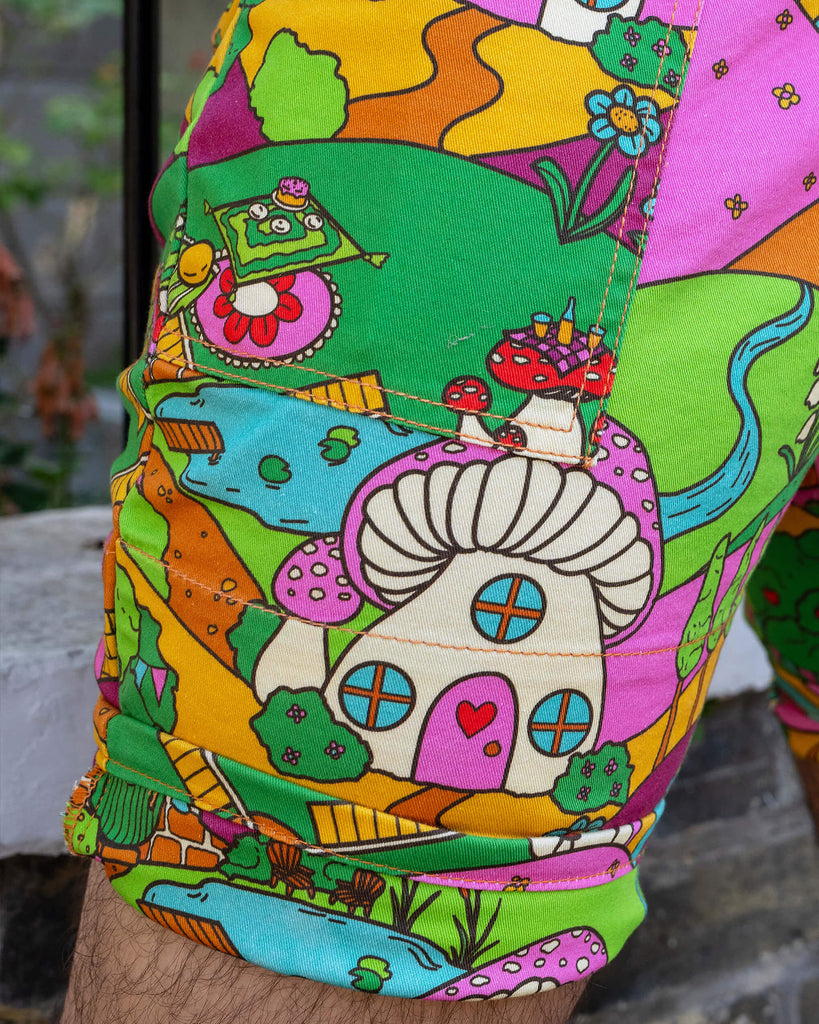 Close up of the Run & Fly x The Mushroom Babes Hills Stretch Twill Dungaree Shorts worn by a masc non binary person. Featuring the matching turn up detailing and side pocket.  The dungashorts are green, pink and orange hills with mushroom houses, flowers, rainbows and trees.