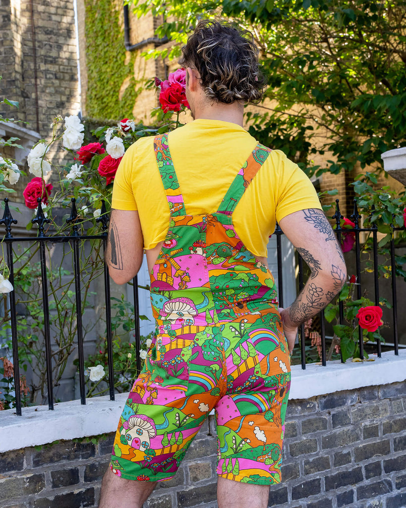 The Run & Fly x The Mushroom Babes Hills Stretch Twill Dungaree Shorts worn by a masc non binary person with a curly brown mullet haircut, a yellow crop top and white trainers. They are stood outside in front of a black railing with foliage and rose bushes behind. They are facing away with one hand on their hip. The dungashorts are green, pink and orange hills with mushroom houses, flowers, rainbows and trees.