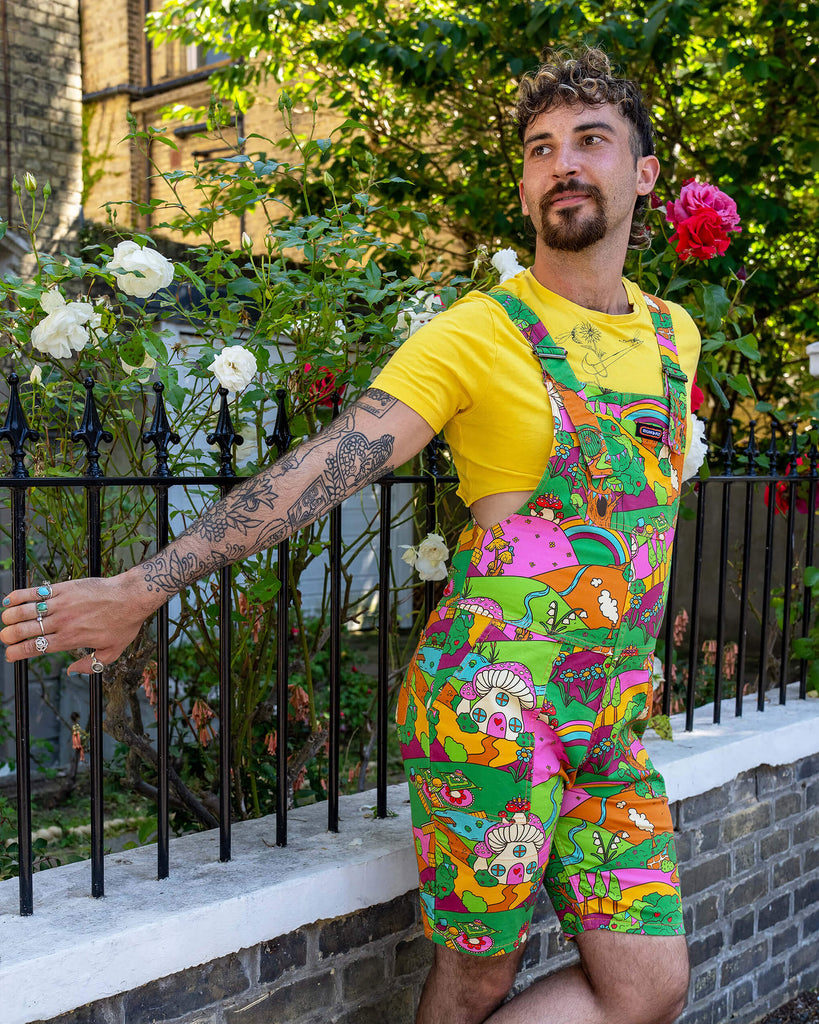 The Run & Fly x The Mushroom Babes Hills Stretch Twill Dungaree Shorts worn by a masc non binary person with a curly brown mullet haircut, a yellow crop top and white trainers. They are stood outside in front of a black railing with foliage and rose bushes behind. They are facing forward leaning off the railing with one leg crossed in front of the other smiling looking left. The dungashorts are green, pink and orange hills with mushroom houses, flowers, rainbows and trees.