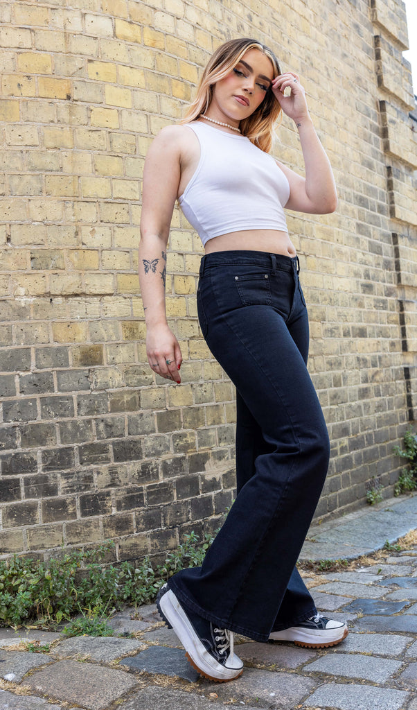 Melia a white femme person wit dyed caramel coloured hair is smiling wearing a white vest and black hippy flares by Run & Fly stood in a mews in Hove