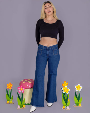 Shannon is wearing Stonewash Blue Stretch Denim Bell Bottom 90's Wide Leg Hippy Flares with a black cropped long sleeve top and white boots. She is stood in front of a grey backdrop with a toad stool and flower decorations next to her. She is posing toward the camera with her hands behind her back.