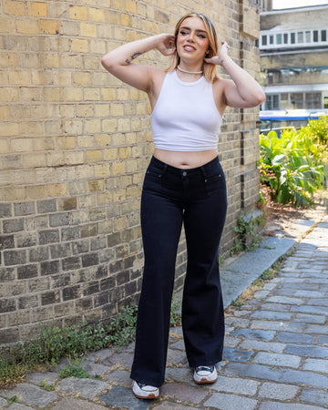 Melia a white femme person with dyed caramel coloured hair is smiling wearing a white vest and black hippy flares by Run & Fly stood in a mews in Hove