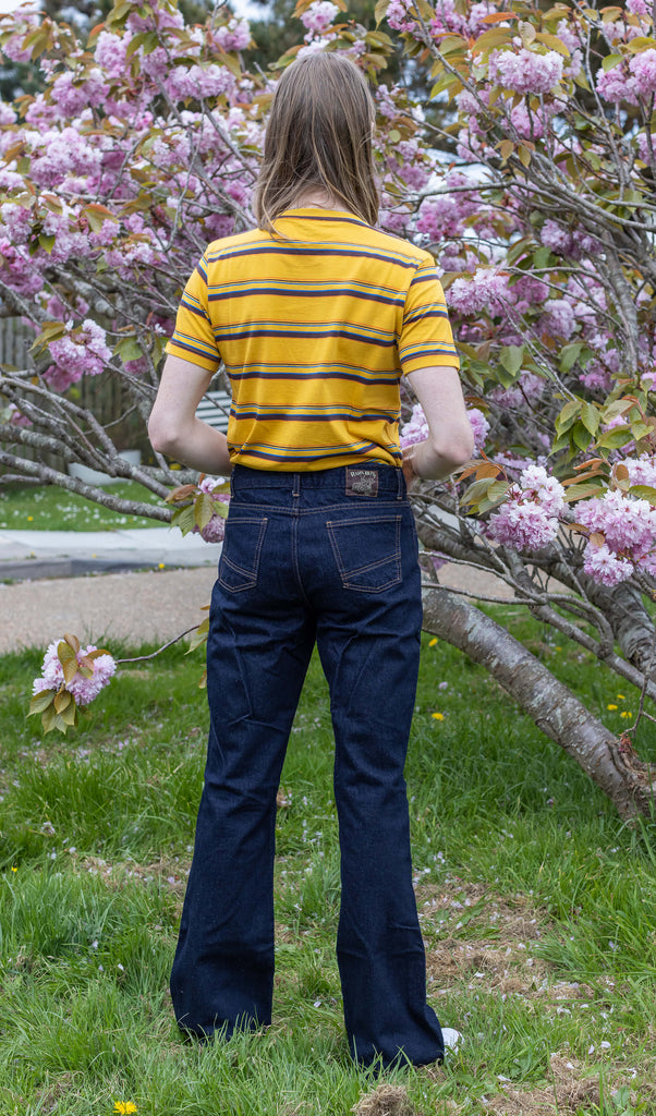 Jack a blonde male model with shoulder length hair is looking at camera infant of a blossom tree wearing a mustard striped tee and indigo blue flares. 
