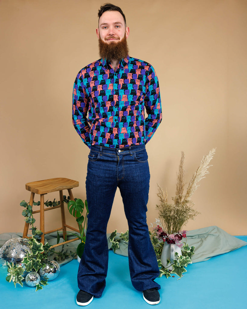 male model wearing Indigo Blue Mid-Low Rise Bell Bottom Super Flare paired with Cat Chorus Print Long Sleeve Shirt. He is stood in a studio in Hove smiling toward the camera with his hands behind his back. 