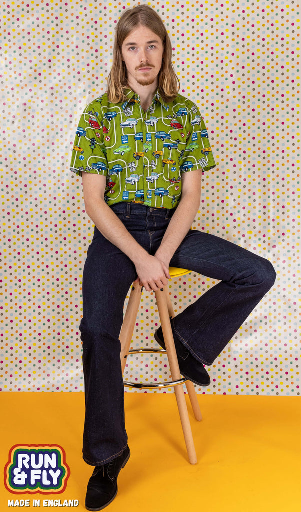 Model is sat on a stool in front of a multicoloured dotted studio background wearing a green race car short sleeve shirt and the denim retro boot cut flare jeans with smart black shoes. They are sat facing the camera with one leg resting on the stool footrest, both hands are falling into their lap.