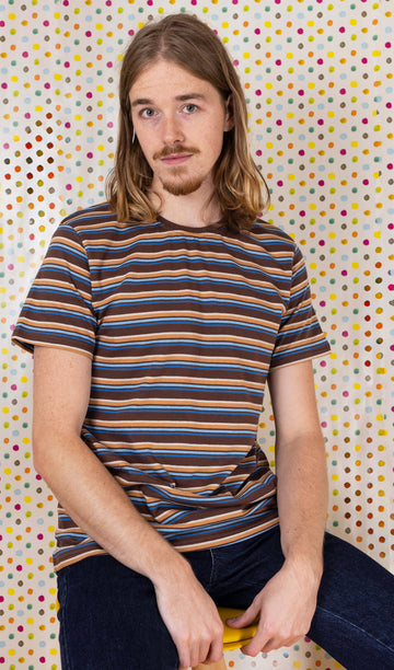 Model is sat in front of a multicoloured dot studio background wearing the brown stripe retro t-shirt with denim jeans. They are sat facing the camera with both hands resting by their sides and smiling.