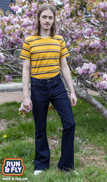 Jack a blonde male model with shoulder length hair is looking at camera infant of a blossom tree wearing a mustard striped tee and indigo blue flares. 