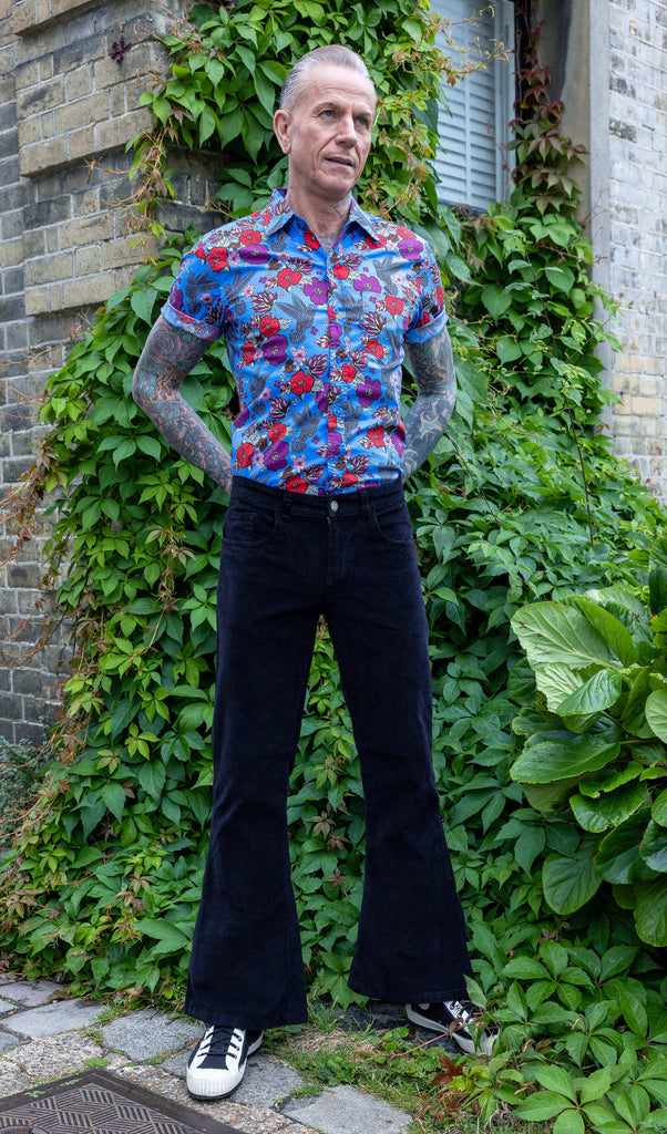 James is wearing Black Corduroy Bell Bottom Flares, paired with Hummingbird Print Short Sleeve Shirt and white trainers. He is posing with hands on his hips.