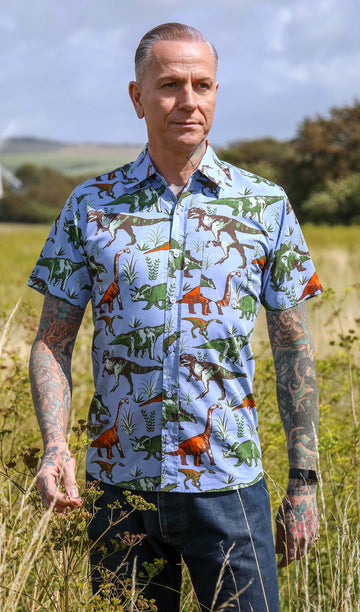 James a white male with silver hair and tattoos in his 50's is looking off to the side in a field wearing a blue dinosaur shirt and jeans