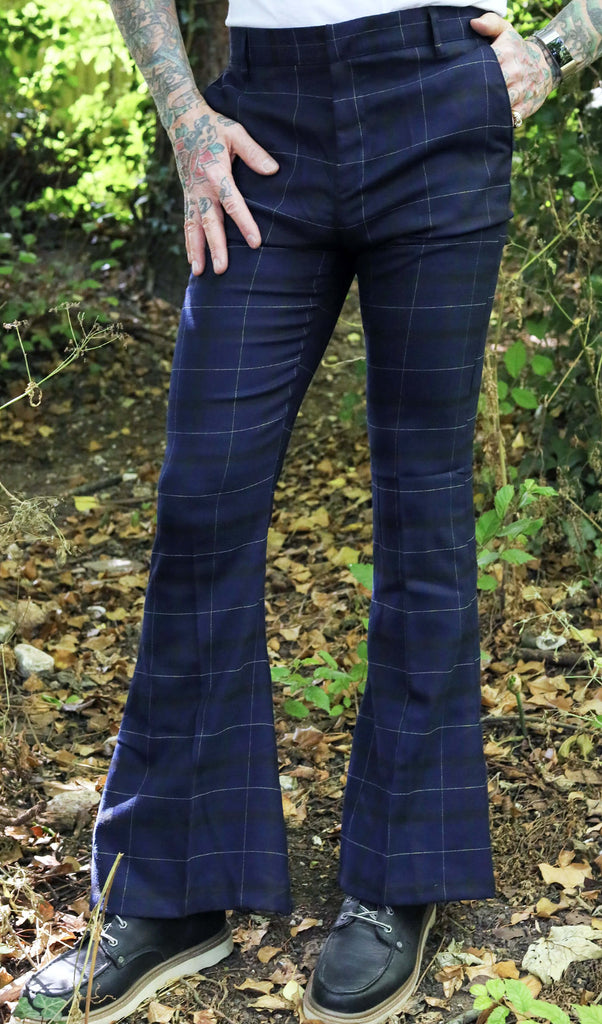 Model is stood outside in a wooded area wearing the blue tartan stretch plaid bell bottom trousers in dark blue. Model is posing facing towards the camera with one leg to the side, one hand is in his pocket whilst the other is resting by his side. Photo is cropped from the waist down. He has paired the trousers with a white t-shirt and brown boat shoes.
