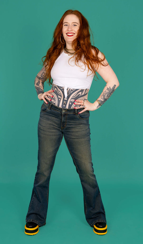 Isobella a tattooed model with long ginger hair is wearing Unisex Distressed Denim Bellbottom Flares Mid Rise Jeans paired with a white crop top and black and yellow boots. The model is stood smiling at the camera with her hands on her hips and the background of the photo is green. 