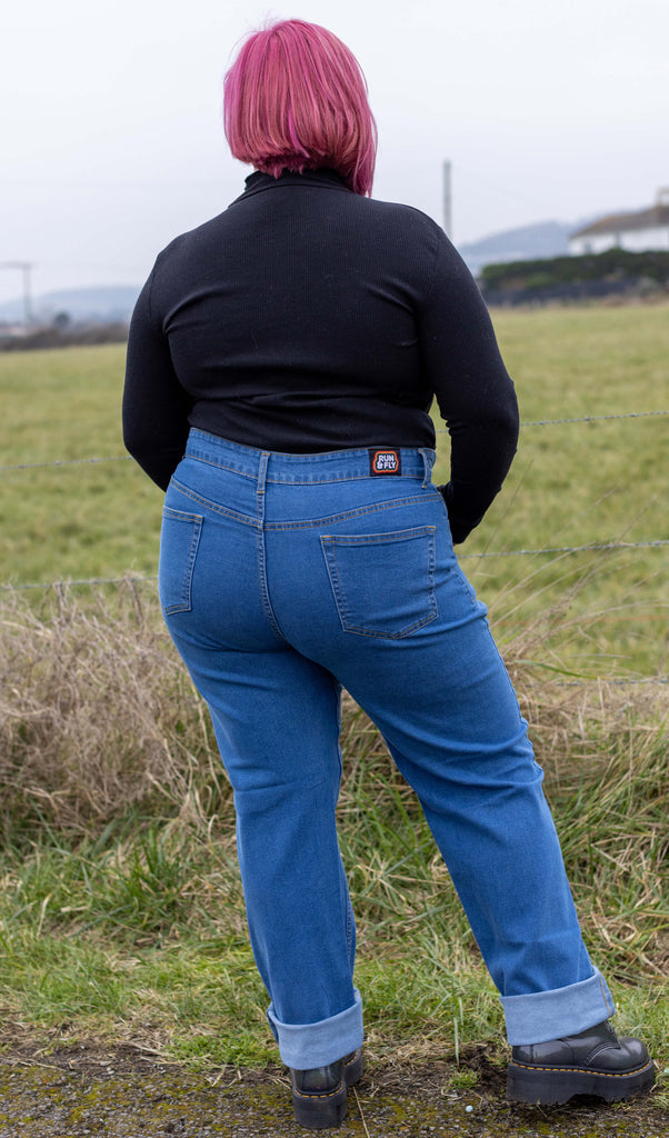 Model is stood in front of a grass field wearing the 90s stonewashed straight leg high waisted jeans with a long sleeve black top and grey boots. They are facing away from the camera with both arms folded in front of them whilst leaning more on one leg to the left.