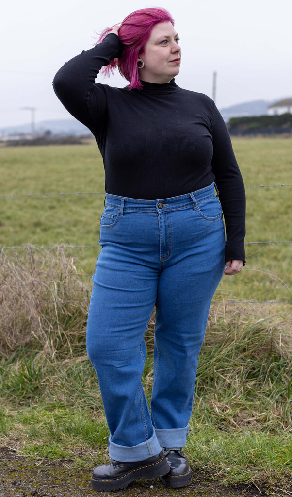 Model is stood in front of a grass field wearing the 90s stonewashed straight leg high waisted jeans with a long sleeve black top and grey boots. They are facing forward posing with one leg crossed in front of the other and one hand running through their pink hair whilst looking off to the right.