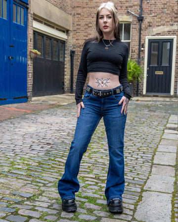 Front-facing view of the Run & Fly Stone Wash Blue Distress Bell Bottom Flares styled with a studded belt and black crop top, worn by a model standing on a cobbled street.
