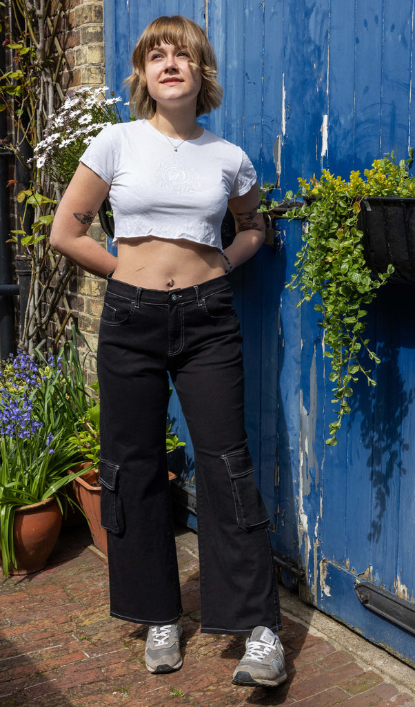 The Black Contrast Stitch Wide Leg Stretch Cargo Trousers modelled by a femme person with short blonde hair, a white crop top baby tee and grey trainers. She is stood posing in front of a blue wooden door and plant pots outside leaning on her left leg, both hands resting behind her hips smiling looking off to the left. The black cargo trousers have white contrast stitching with two front flap pockets on the lower legs.