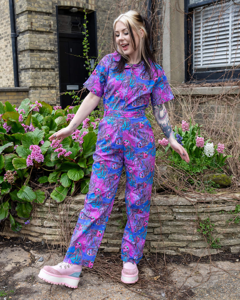 Model smiling and posing in the Run & Fly Jellyfish Stretch Jumpsuit, showing the full-length straight leg fit, bright multicolour jellyfish print, and short-sleeve top half.