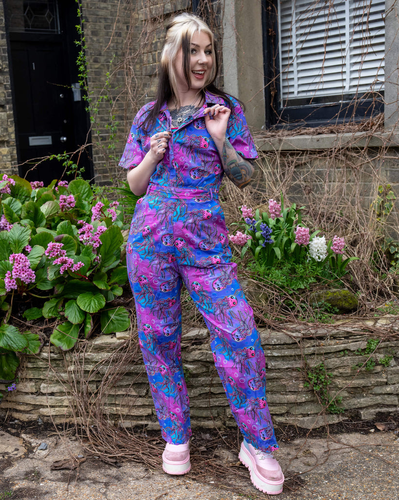 Front view of the Run & Fly Jellyfish Stretch Jumpsuit, with the model smiling and unzipping the front, styled with pastel pink platform boots and spring flowers in the background.