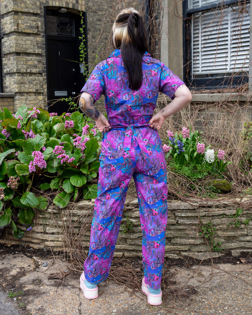 Full back view of the Run & Fly Jellyfish Stretch Jumpsuit worn by a model with hands on hips, surrounded by spring flowers and stone walls, highlighting the colourful all-over print.