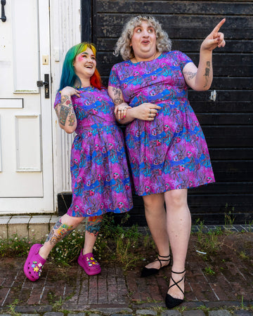 Two women standing close together and laughing, both dressed in the Run & Fly Jellyfish stretch belted tea dress with pockets. The vibrant purple and blue dresses have a jellyfish pattern. The woman on the left has rainbow-coloured hair and colourful tattoos, while the woman on the right has curly blonde hair and also displays tattoos. The brand Run & Fly is known for its ethical and slow fashion approach.