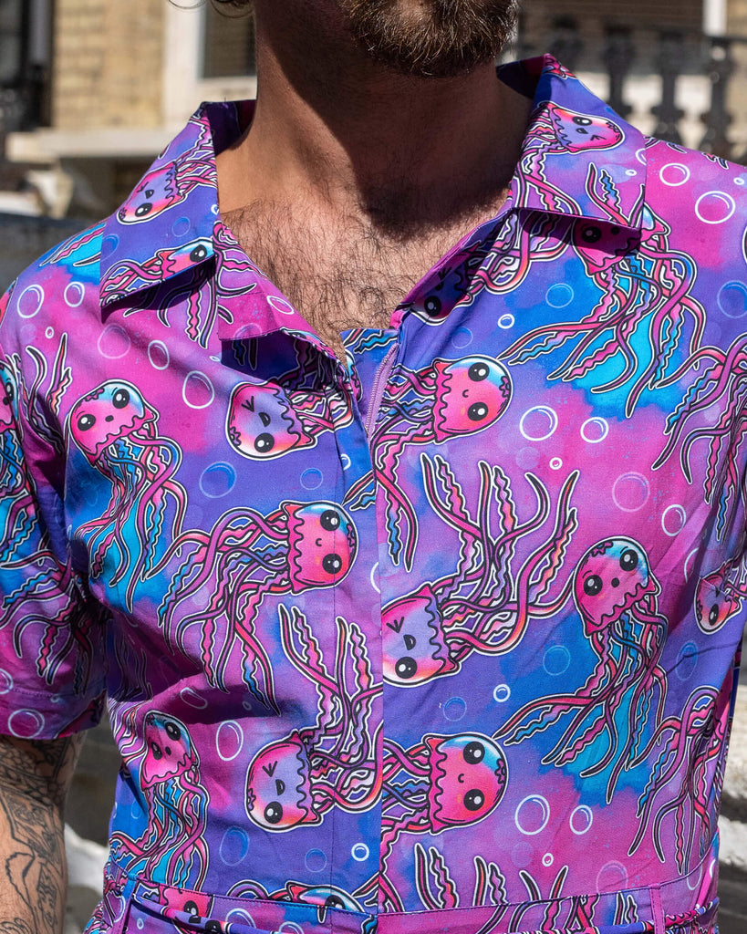 Close up of the Jellyfish Jumpsuit worn by a masc non binary person with a curly brown mullet outside on a pavement with tall ornate houses, white concrete and brick walls and purple flowers growing along the bottom. They are facing forward with both hands resting in the side pockets highlighting the zip up front and belt tie detailing. The blue, pink and purple base print features various smiling pink, blue and purple jellyfish and white bubble outlines.