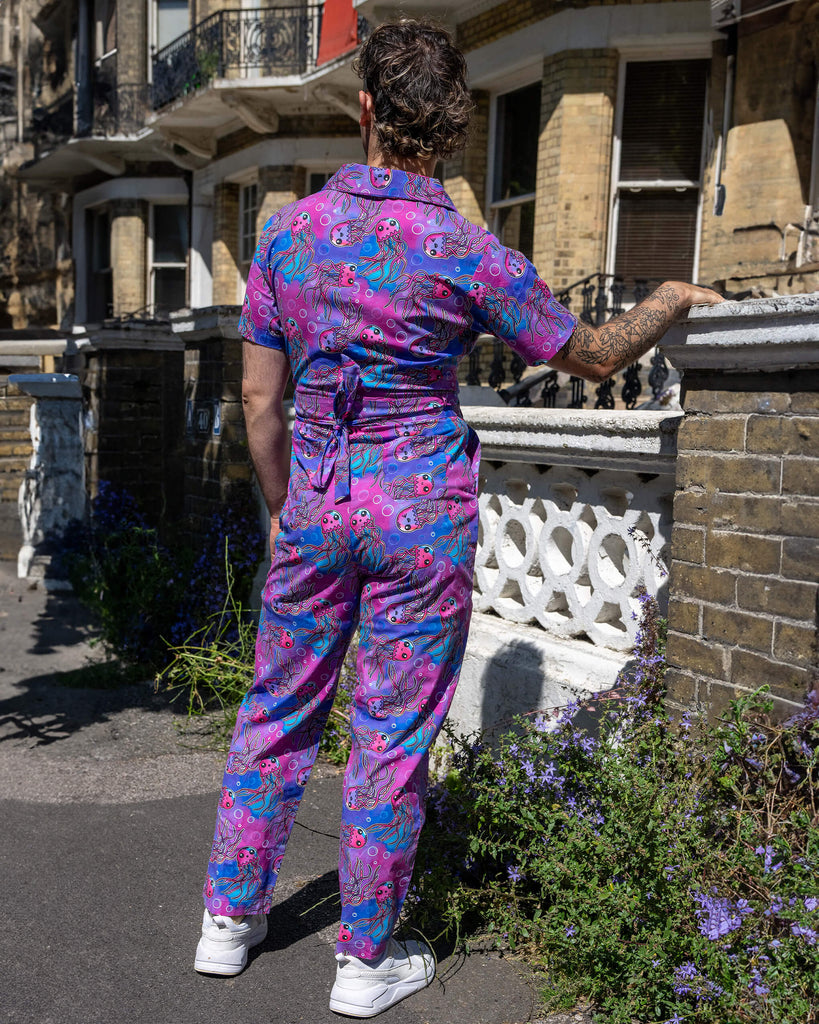 The Jellyfish Jumpsuit worn by a masc non binary person with a curly brown mullet and white trainers outside on a pavement with tall ornate houses, white concrete and brick walls and purple flowers growing along the bottom. They are facing away leaning one hand on the wall. The blue, pink and purple base print features various smiling pink, blue and purple jellyfish and white bubble outlines.