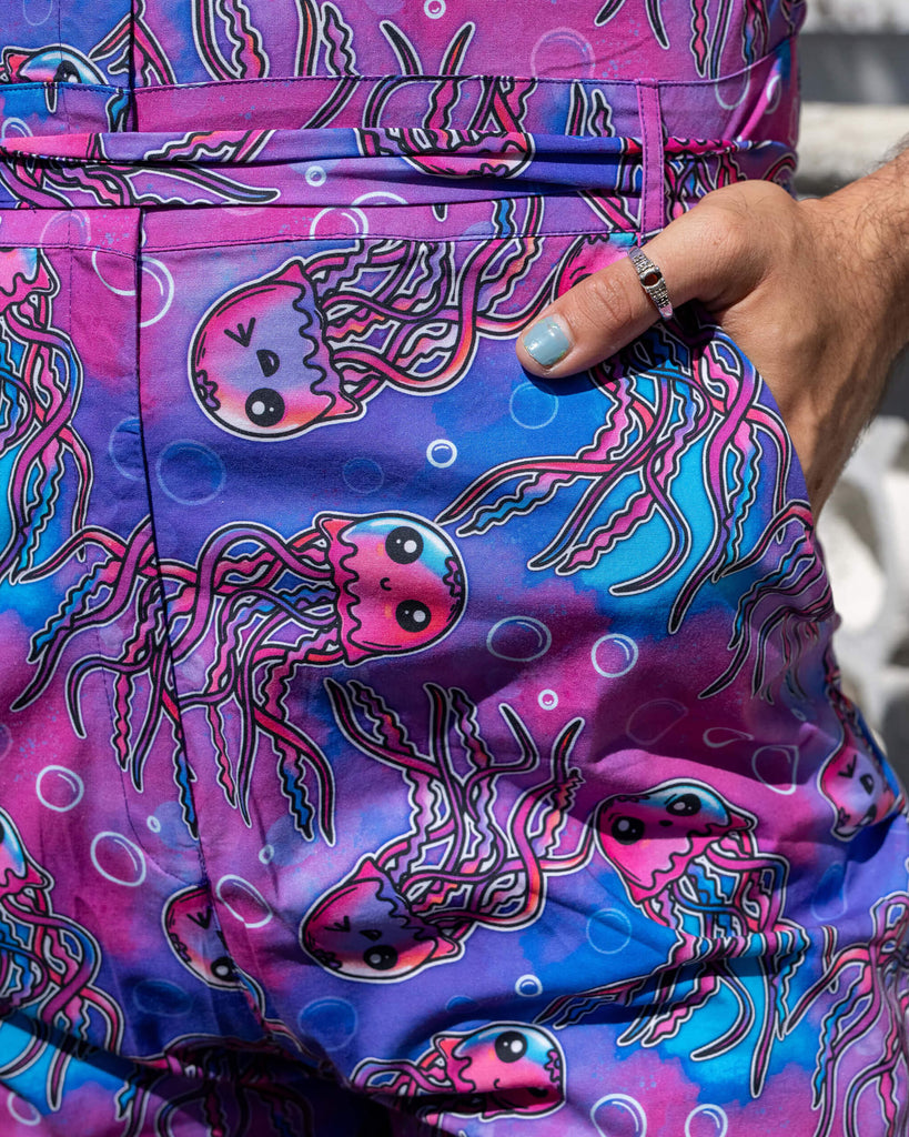 Close up of the Jellyfish Jumpsuit worn by a masc non binary person with a curly brown mullet outside on a pavement with tall ornate houses, white concrete and brick walls and purple flowers growing along the bottom. They are facing forward with one hand in the pocket highlighting the belt tie detailing and the print. The blue, pink and purple base print features various smiling pink, blue and purple jellyfish and white bubble outlines.