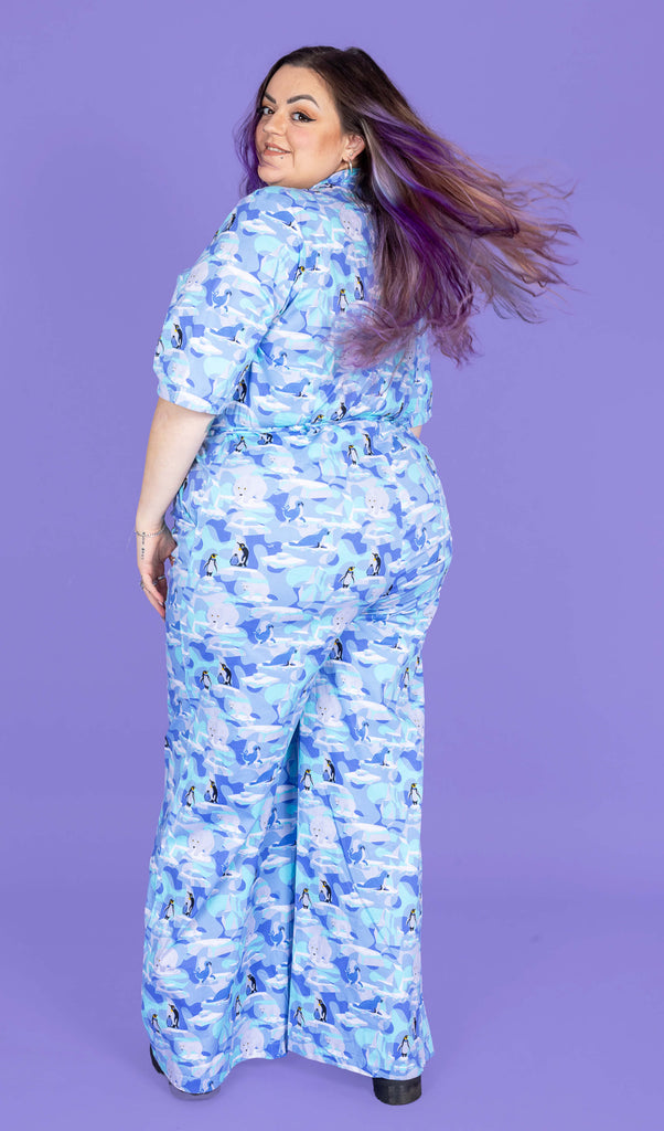 a model with purple hair wearing Winter Friends Stretch Jumpsuit paired with black boots. The jumpsuit is a light blue colour with ice burgs, seals, polar bears and penguins on. Model is standing with their back to the camera and looking back over their shoulder with their hair swishing. The background of the photo is purple.