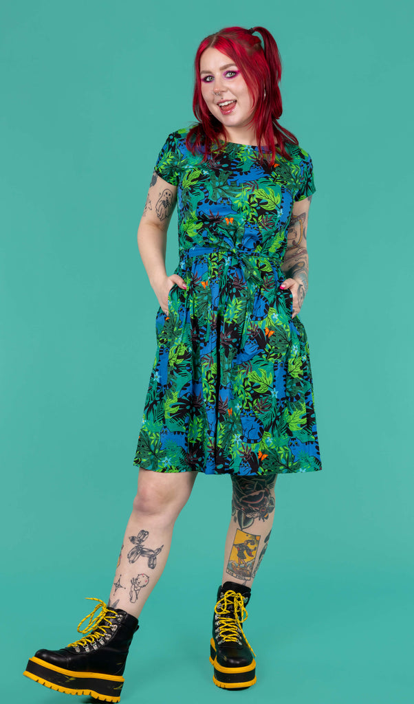 The Jungle Cats Stretch Belted Tea Dress with Pockets being worn by Flo on a teal background. Print features various green, brown and black foliage with orange butterflies and dark blue jumping tabby cats, the dress is short sleeve with a belt tie and pockets. Flo has red hair in bunches, neon pink makeup and tattoos with yellow and black platform boots. She is facing forward smiling leaning back on one leg with both hands in the dress pockets.