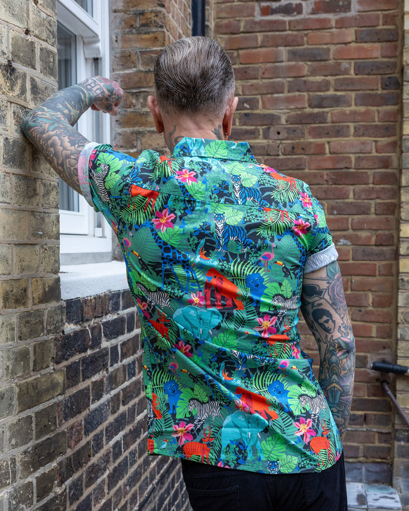 James is wearing Jungle Stretch Short Sleeve Shirt, paired with black trousers. The collared shirt is a forest green colour with all over print of various jungle animals and plants in black, white, red, pink, yellow and blue. James is leaning against a wall with back to camera to show back of shirt.